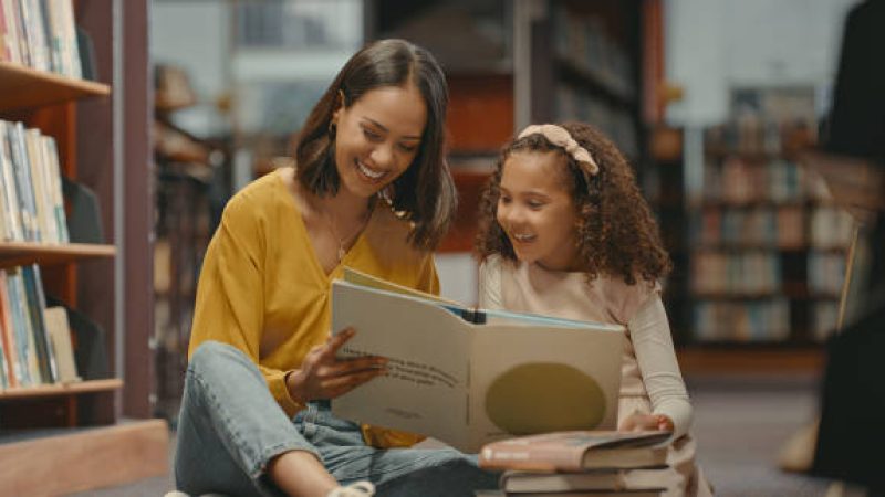 Teacher helping a young student with her homework in the library after school. Two females are reading a book together in the bookstore. They are doing research for a project